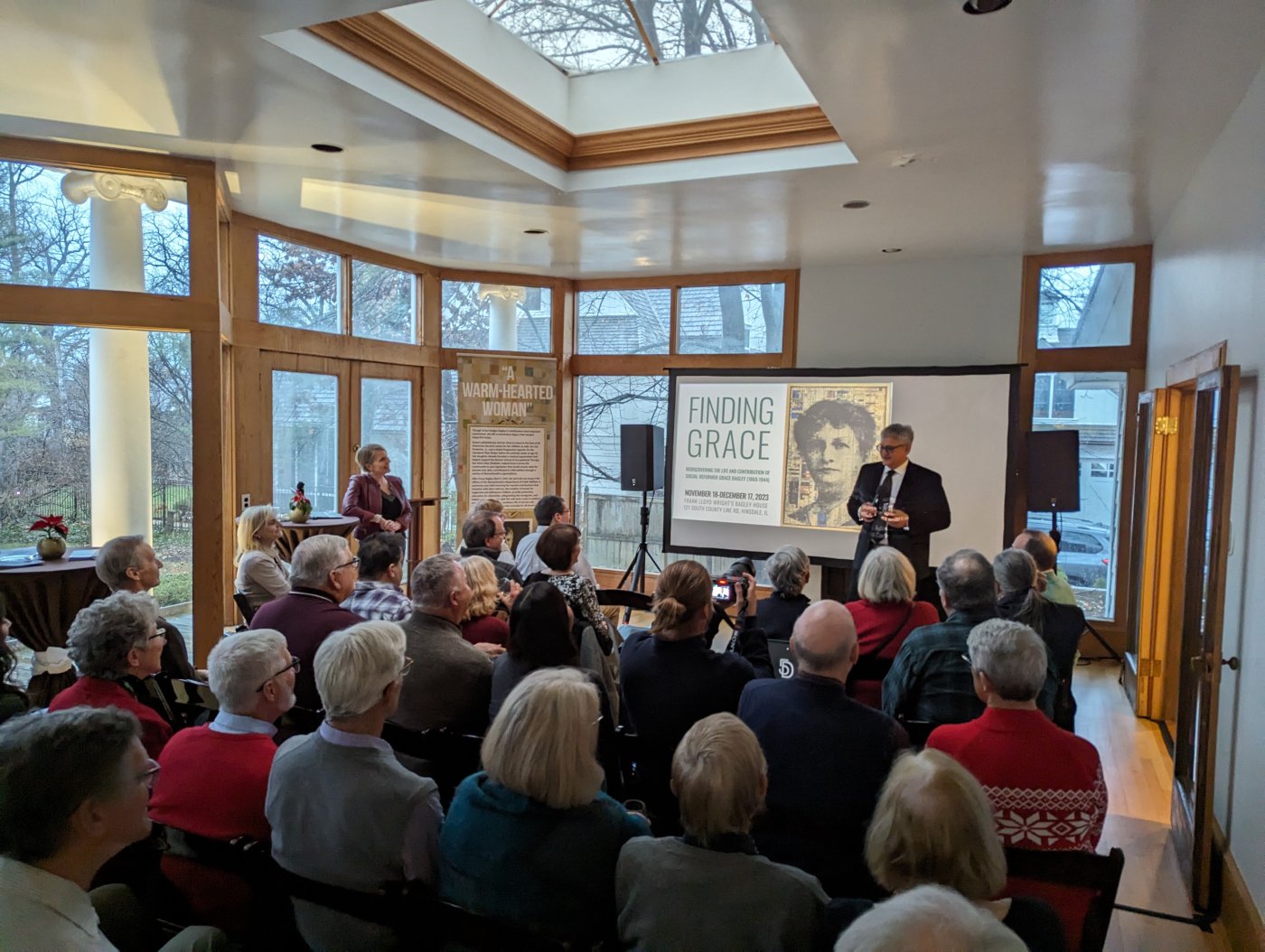 A crowd of people seated in the window-lined room at the back of the Bagley House, being addressed by a man standing at the front with a screen showing the title slide for the "Finding Grace" exhibit