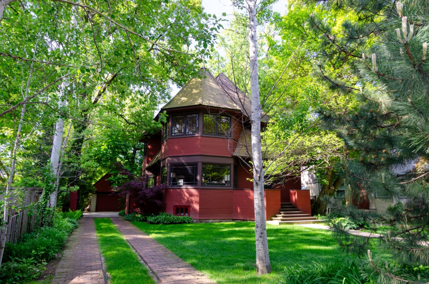 Front exterior view of Parker House with driveway, green lawn, trees all around, and red two-story wood house with windows on both levels of an octagonal turret on the left under a pitched roof, and stairs leading up to a porch on the right