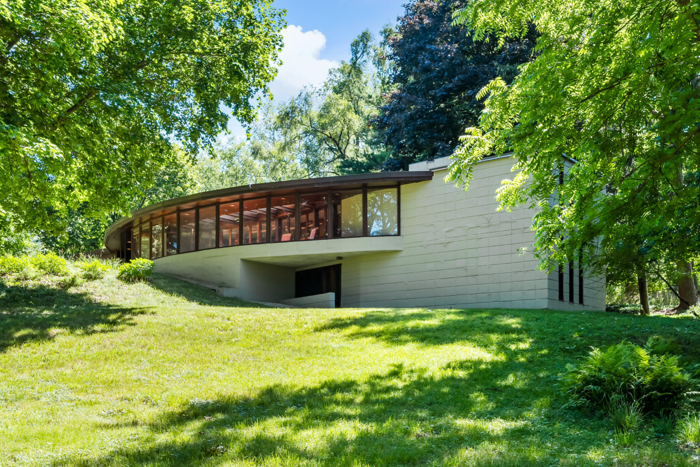 Exterior view looking up hill across green grass towards a concrete block house wall, with a curved wall of windows emanating to the left at the crest of the hill.