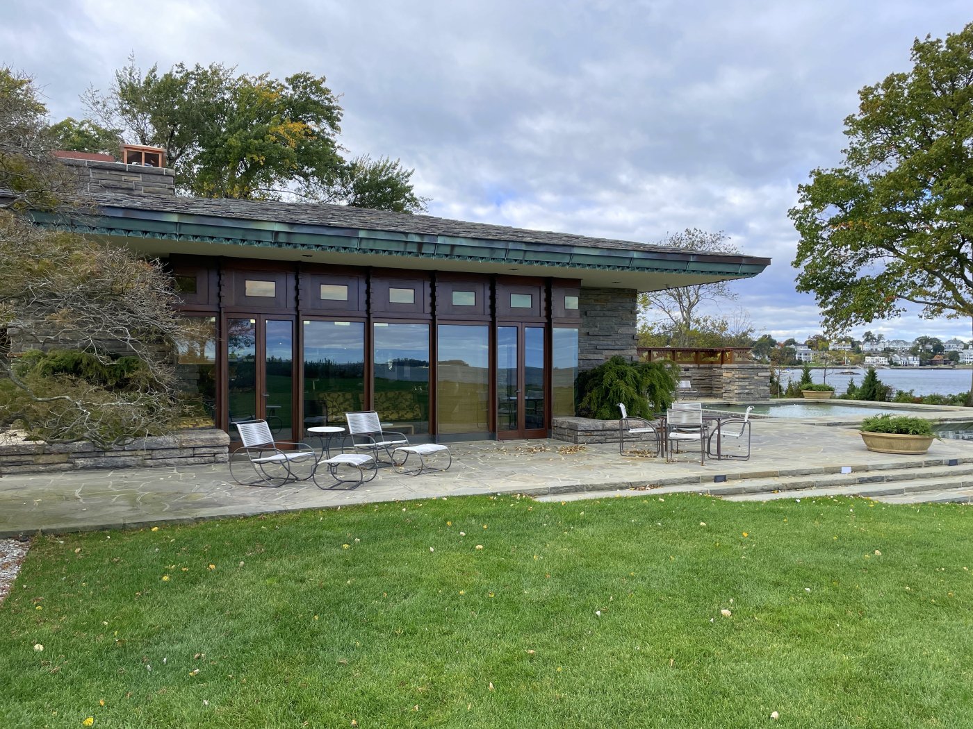 Exterior of house showing low-angle sloped roof with copper fascia, window wall of mostly glass doors with some wood, a stone patio, and the waters of Long Island Sound in the distance to the right. Green lawn in foreground.