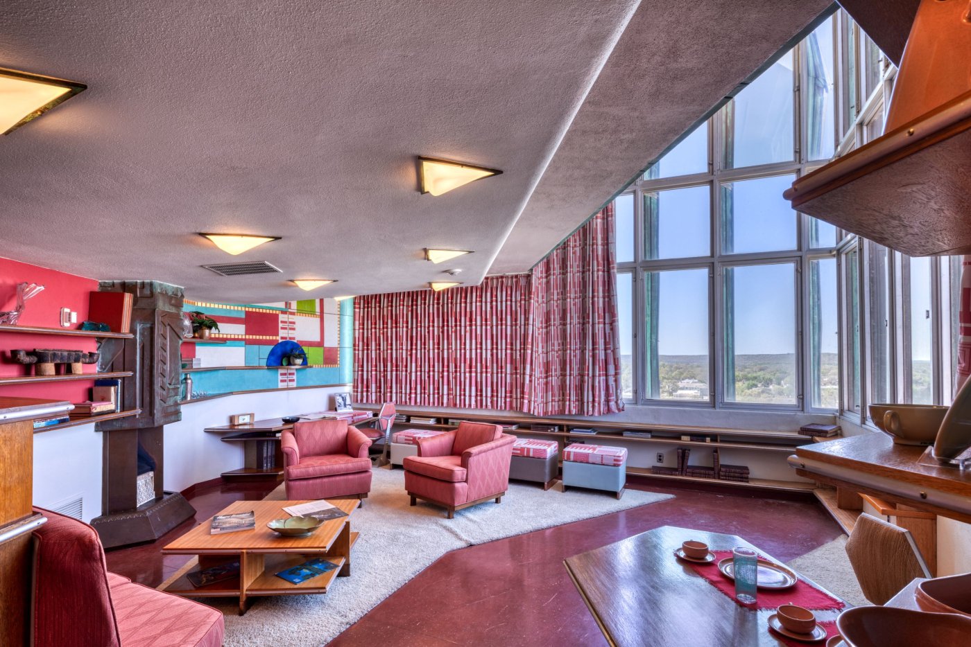 Interior space at Price Tower showing a low ceiling to the left with triangular light fixtures in it, a fireplace with copper hood with geometic designs and built-in shelves on the right wall, furniture on a white rug on top of the red floor, and opening up to a much taller wall of windows to the right