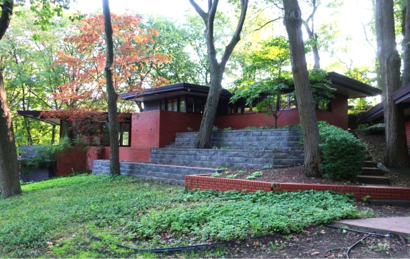 Photo of exterior of brick house built atop stone terraces on a sloped and wooded site, with bands of horizontal windows and a flat roof with wide overhanging eaves.