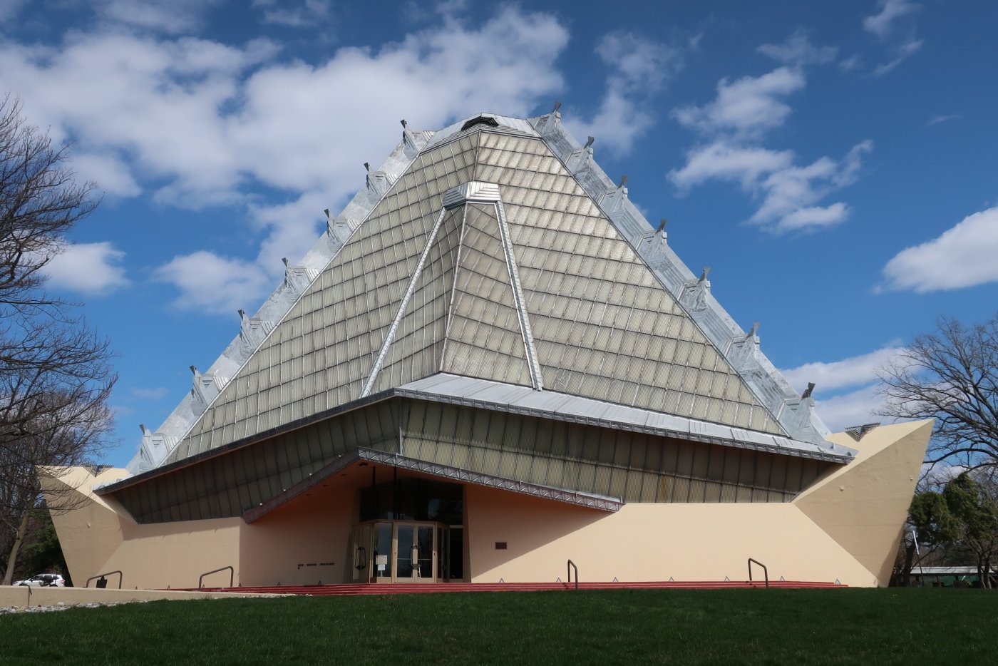 Photo of exterior of synagogue showing dramatic pyramidal roof framed in metal with opaque glass panels, resting on an angular cream-colored concrete base.