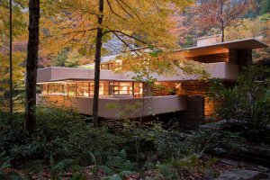 Photo looking through trees and shrubs with some fall colors towards the house of Fallingwater, with warm light emanating from inside through windows on two different levels, heavy horizontal concrete balcony walls, and stone walls catching the light in a few different places.