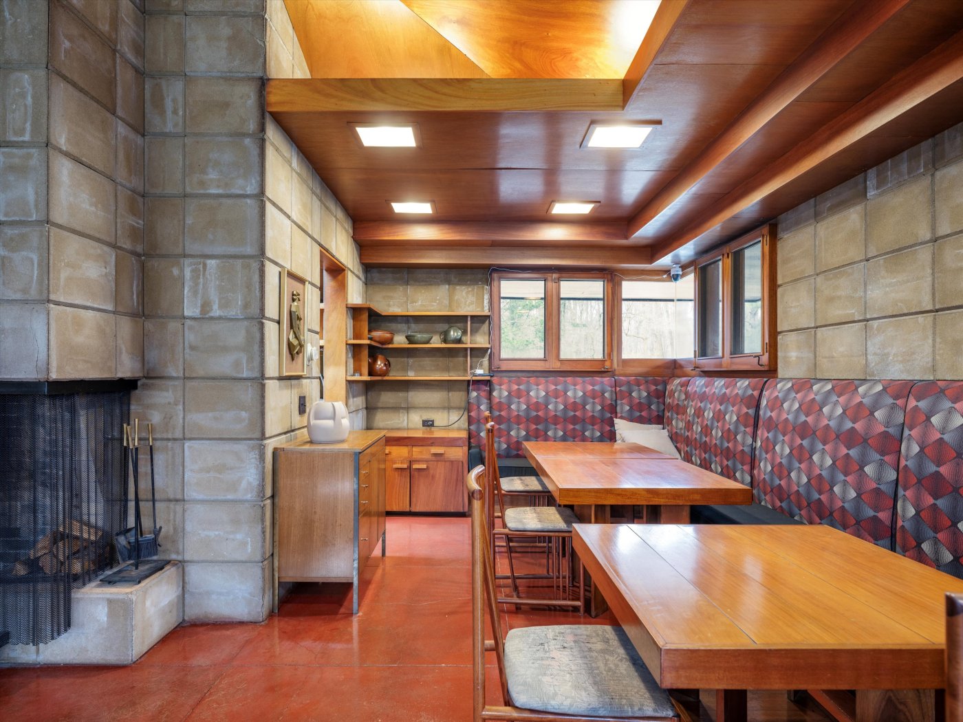 Interior of house showing fireplace and concrete block wall to left, wood ceiling with recessed lights above, red concrete floor, and wood built-ins to the right.